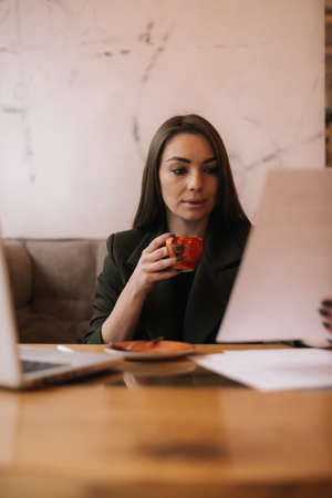 Beautiful young business woman studying paper documents in cafe sitting at table with laptop. Lady enjoys delicious coffee while working. Student girl sitting in cafe with cup of coffee.の写真素材