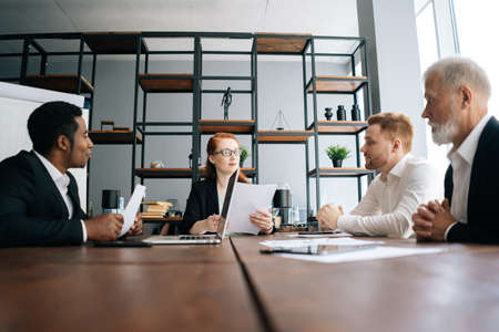 Motivated business woman leader holds meeting with employees at office desk, shooting from below. African American businessman with laptop listening question and answering to boss.の写真素材