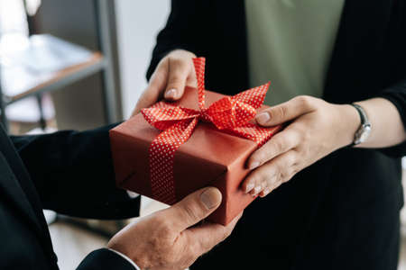 Close-up view of hands of unrecognizable woman giving red gift box tied to bow handed to man. Giving gifts during the Christmas, Happy New Year and Happy Birthday at office.の写真素材