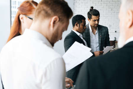 Multicultural coworkers examining financial data on paper document at office room. Young creative diverse multi-ethnic team people brainstorm on project strategy share development ideas near window.の写真素材