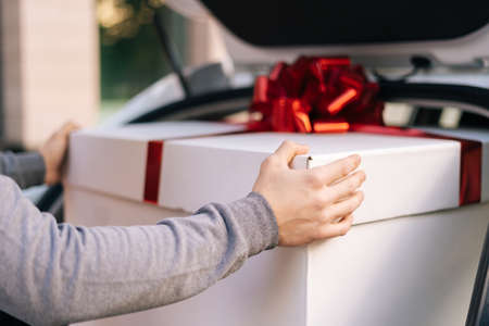 Close-up view of delivery man standing near car preparing to take parcel out of trunk of car and deliver it to customer. Young male makes surprise for girlfriend or wife.の写真素材