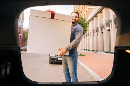 Portrait of smiling delivery man holding large festive box with red bow near car outdoors city...
