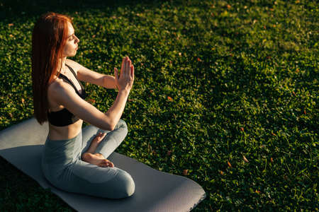 High-angle shot of young lady with closed eyes is meditating in lotus position sitting on yoga mat,...