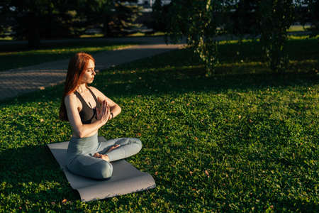 Young red-haired woman with closed eyes wearing sportswear sitting in lotus position doing yoga...