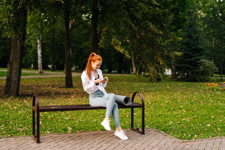 Red-haired joyful young woman wearing sportswear watching video on smartphone sitting bench at city park. Attractive female walking chatting with friends using cellphone outdoors.の写真素材