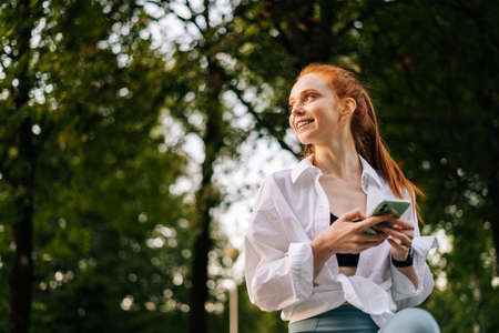Low-angle shot of cheerful young woman browsing internet on smartphone, looking away. Portrait of...