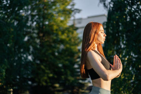 Side view of attractive redhead young woman with closed eyes practicing yoga and meditation performing namaste pose. Calm female making Namaste gesture outside in city park in evening at sunset.の写真素材