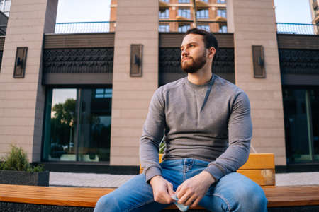 Low-angle shot of upset young man sitting on urban bench at empty European city street. Relaxed handsome male having resting outdoors on empty street. City on quarantine. Commercial business shutdown.の写真素材