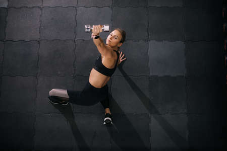 Top down view of strong young woman with perfect athletic body lying on floor working out lifting dumbbell during sport workout training at modern fitness gym with dark interior.の写真素材