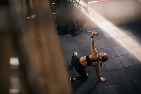 Top down view of strong young woman with perfect athletic body lying on floor working out lifting dumbbell during sport workout training at modern fitness gym with dark interior.の写真素材