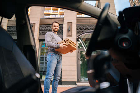 Bearded handsome Caucasian delivery man standing at city street and holding carton boxes with hot...