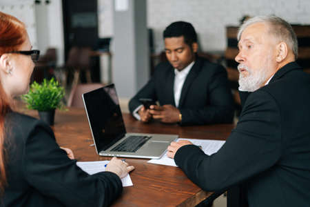 Skilled mature adult male businessman having meeting with young diverse multi-ethnic business colleagues in modern office. Multiethnic business team of businessmen people discussing new project.の写真素材
