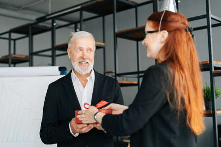 Business woman giving red gift box to smiling mature adult male leader during the Christmas, Happy New Year and Happy Birthday at office. Happy business lady congratulate to colleague.の写真素材