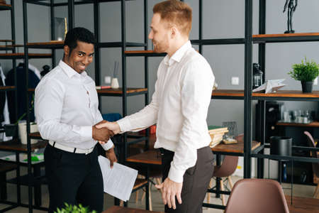Cheerful diverse business partners shaking hands in agreement standing in office hall. African American and Caucasian male colleagues wearing formalwear shaking hands and discussing businessの写真素材