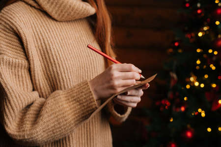 Close-up hands of unrecognizable young woman writing Christmas letter to Santa Claus at cozy dark living room. Xmas tree baubles and tinsel lights blinking at New Year Eve on blur bokeh background.の写真素材