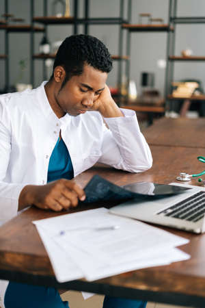 Side view of ponder African American black male doctor in white coat analyzing history disease of patient using MRI brain head scan, working on laptop. Concept of medicine and health care.の写真素材