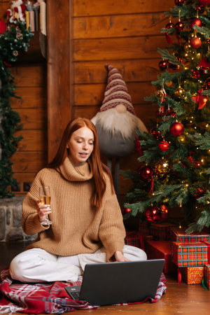 Charming red-haired young woman talking on laptop to webcamera sending online congratulations holding glass with champagne, sitting on floor background of Christmas tree at cozy living room.の写真素材