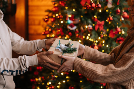 Close-up hands of African American man giving Christmas gift to Caucasian girlfriend on background xmas tree with celebration lights at cozy room with festive interior. Couple enjoying celebration.の写真素材