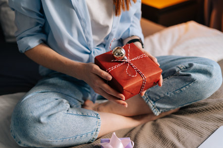 Close-up hands of unrecognizable woman holding gift box with holiday present during online communication using laptop. Happy lady sitting on lotus position at bed at bedroom and having video chat.の写真素材