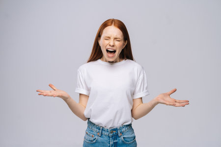 Angry frustrated young woman wearing T-shirt and denim pants screaming out loud on isolated white background. Pretty lady model with red hair emotionally showing facial expressions.の写真素材