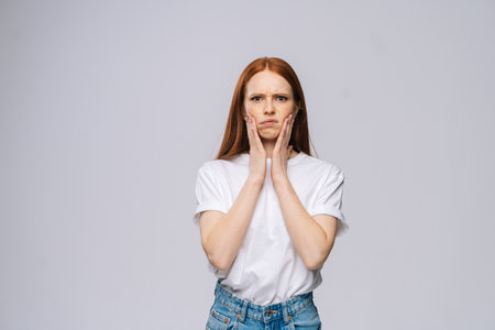 Disgruntled charming young woman wearing T-shirt and denim pants holding hands on face on isolated...