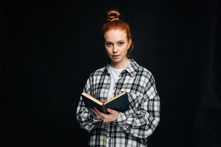 Attractive young woman college student holding opened book and looking at camera on black isolated background. Pretty redhead lady model emotionally showing facial expressions in studio, copyspace.の写真素材