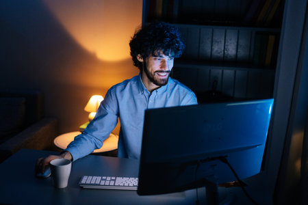 Top view of happy smiling bearded young man looking intently at computer monitor screen while working on at late night. Concept of remote working, distance learning, Concept of remote working.の写真素材
