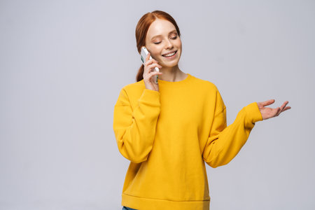 Smiling young woman wearing stylish yellow sweater talking by mobile phone on isolated white...