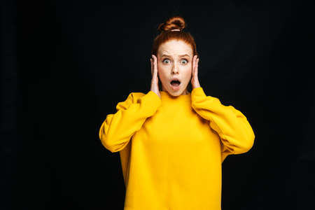 Portrait of shocked afraid young woman looking excited holding mouth opened, hands on face on isolated black background, looking at camera. Pretty redhead lady emotionally showing facial expressions.の写真素材