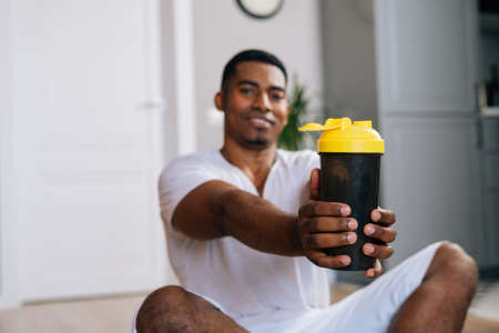 Blurred view of confident muscular African-American man sitting cross-legged relaxing on exercise mat after workout and holding proposing bottle of fresh mineral water at domestic room.の写真素材