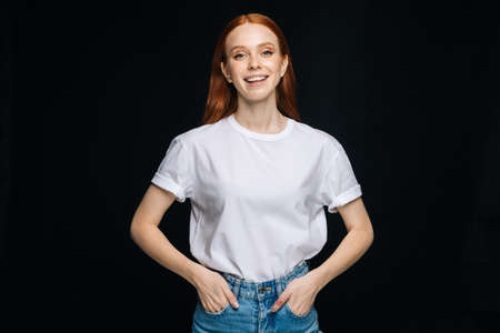 Happy young woman inT-shirt and denim pants posing with hands in pockets on isolated black background, looking at camera. Pretty redhead lady model emotionally showing facial expressions, copy space.の写真素材