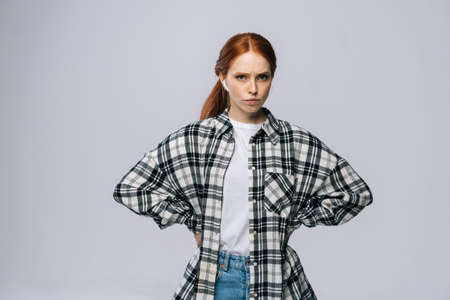 Serious red-haired young woman wearing wireless earphones looking at camera on isolated white...