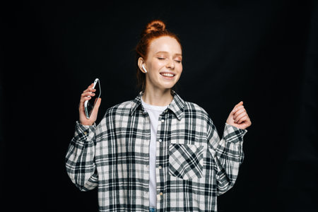 Happy young woman wearing wireless earphones holding phone and dancing listening online music on...