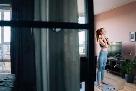 Remote side view of fit woman standing with closed eyes doing stretching exercise for arms and shoulder muscles at home. Concept of sports training red-haired lady during quarantine.の写真素材