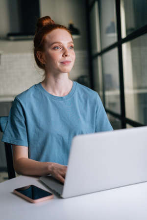 Portrait of thoughtful redhead young woman working typing on laptop computer sitting at table in...