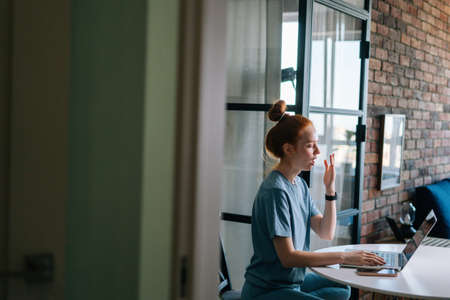 Side view of tired redhead young woman yawning while working on laptop computer sitting at table in cozy room. Concept of remote working from home office during quarantine from COVID-19.の写真素材