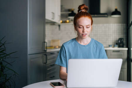 Portrait of pretty redhead young woman working typing on laptop computer sitting at table in kitchen room. Concept of remote working from home office during quarantine from COVID-19.の写真素材