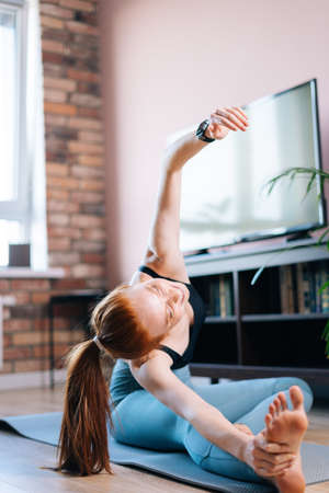 Athletic redhead young woman working out, doing stretching exercise on yoga mat while watching fitness video online on laptop at home. Concept of sports training red-haired lady during quarantine.の写真素材