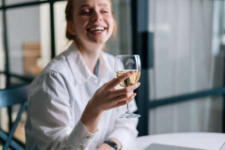 Close-up portrait of laughing redhead young woman holding glass with champagne near face, looking away. Portrait of pretty female with glass of wine.の写真素材