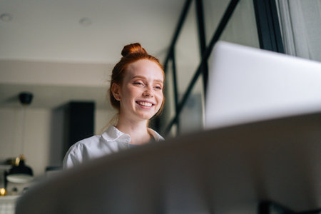 Low-angle view of cheerful redhead young woman having video call via laptop webcam. Concept of leisure activity red-haired female at home.の写真素材