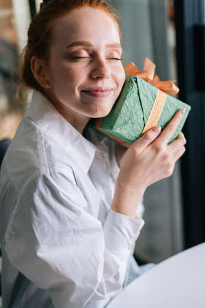 Close-up face of pretty redhead young woman with closed eyes gently cradling surprise gift box to face at home. Concept of leisure activity red-haired female at home during self-isolation.の写真素材