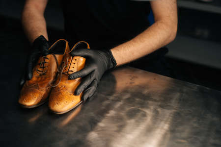 Close-up top view of hands of shoemaker shoemaker in black gloves holding old worn light brown leather shoes on table to be repaired in dark craft shoe shop. Concept of cobbler artisan work.の写真素材