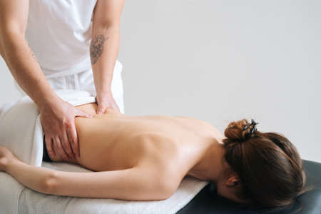 Side view of male masseur massaging lower back of young woman lying on massage table on white...