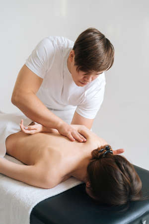 Male masseur massaging back and shoulder blades of female lying on massage table at spa salon. Beautiful young woman with perfect skin getting relaxing massage on white background.の写真素材