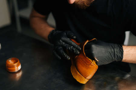 Close-up of unrecognizable shoemaker in black gloves rubbing paint on light brown leather shoes with fingers. Concept of cobbler artisan repairing and restoration work in shoe repair shop.の写真素材