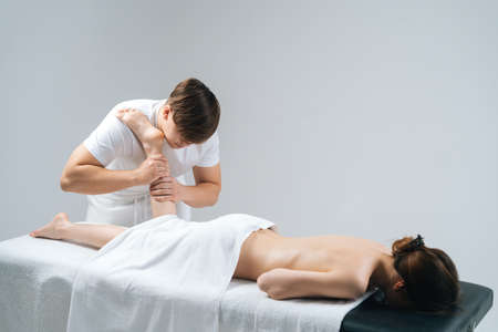 Male masseur with strong hands massaging lower part of leg to young woman lying on massage table on white background. Professional therapist men doing wellness massage shins and legs for female clientの写真素材