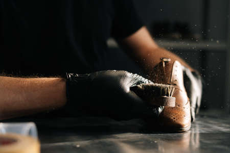 Close-up hands of shoemaker wearing black gloves swipes dust off the shoe brush in dark craft shoe shop. Concept of cobbler artisan repairing and restoration work in shoe repair shop.の写真素材