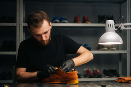 Portrait of shoemaker wearing black latex gloves applying paint on light brown leather shoes and rubbing with fingers. Concept of cobbler artisan repairing and restoration work in shoe repair shop.の写真素材