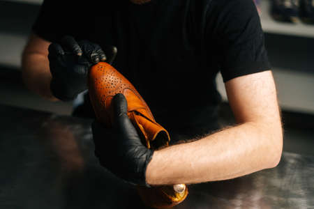 Close-up hands of shoemaker wearing black gloves rubbing paint on toe cup of light brown leather shoes with fingers. Concept of cobbler artisan repairing and restoration work in shoe repair shop.の写真素材
