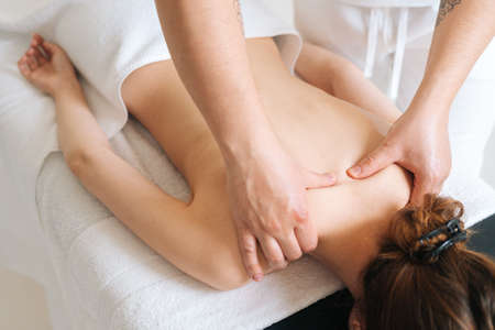 Top view of male masseur with strong hands professionally massaging scapulas and shoulders. Beautiful naked young woman with perfect skin getting back massage at spa salon.の写真素材
